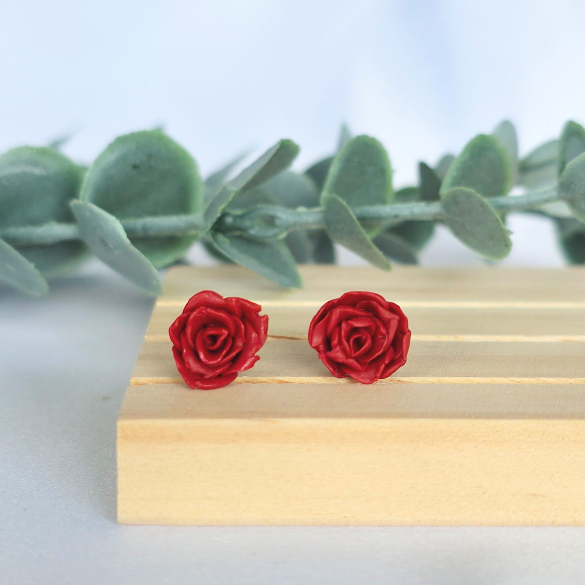 A pair of red rose stud earrings made from polymer clay, displayed on a wooden surface with a greenery backdrop.