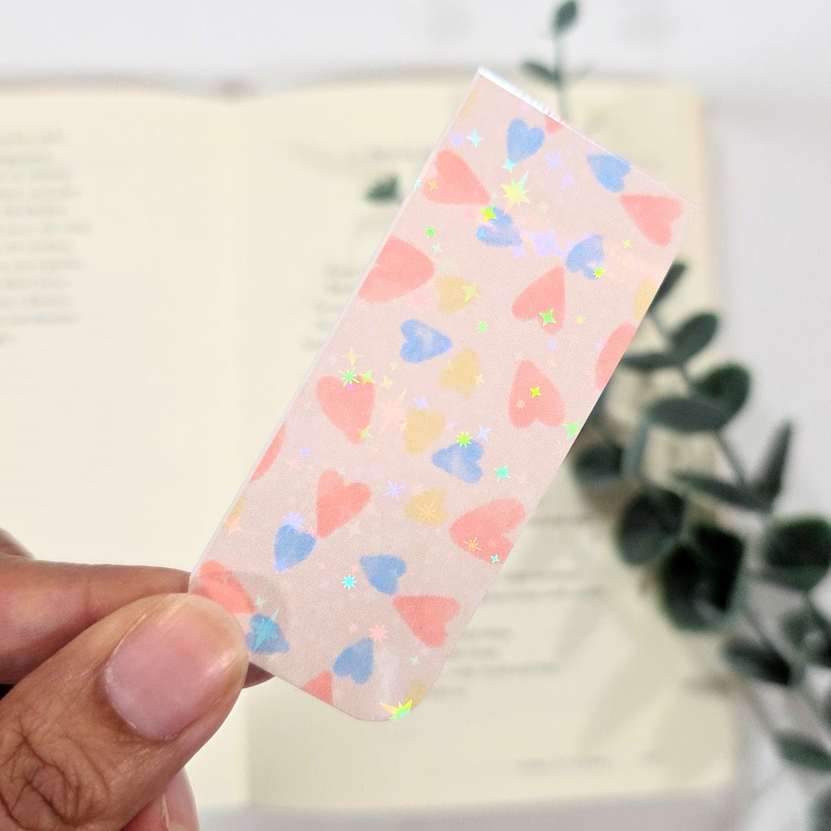 Hand holding a pink bookmark with colorful heart patterns against a blurred indoor background
