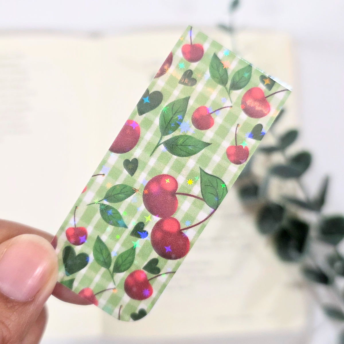 Hand holding a green patterned bookmark with red cherries and green leaves.