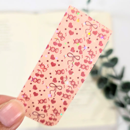 Tan bookmark with heart and 'XOXO' pattern held by a hand against a blurred background