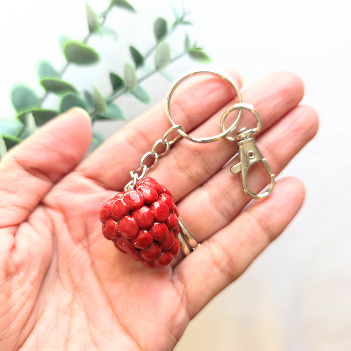 Red raspberry made from polymer clay, keychain on a hand over a white background and greenery.