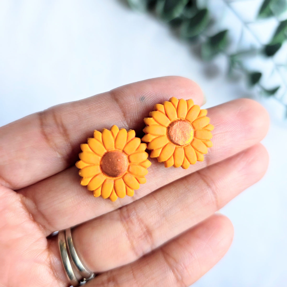Sunflower stud earrings made of polymer clay, held by a hand over a white background and greenery.