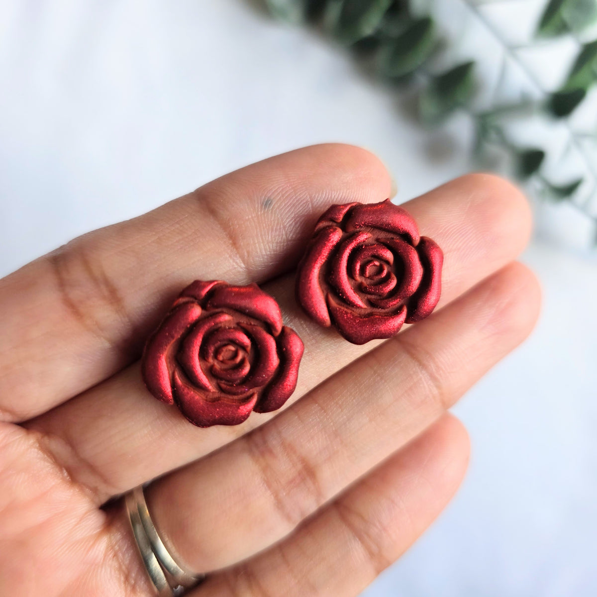 Red Rose polymer clay earring studs held by a hand, and a white background with some greenery.