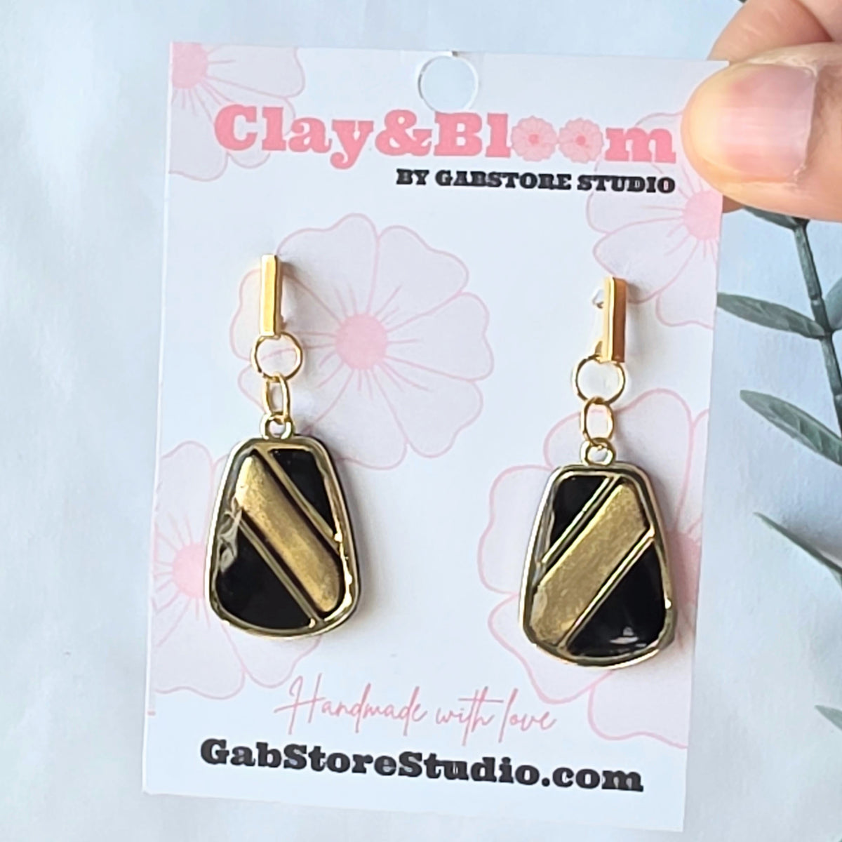 Gold and black earrings on a Clay & Bloom branded card with a hand holding the card.