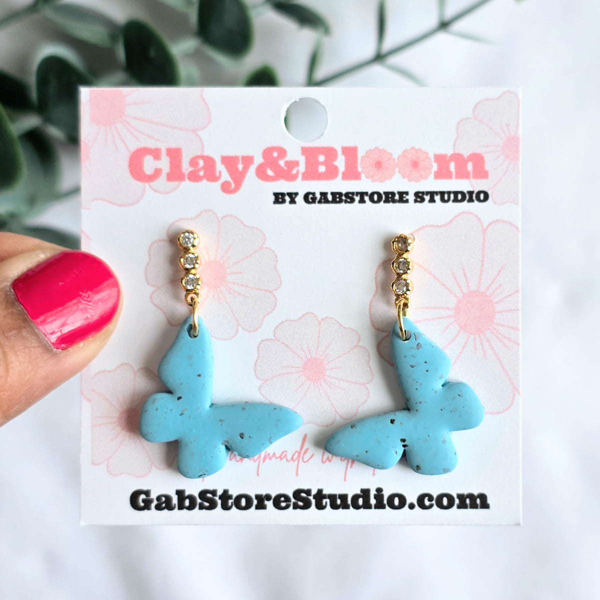 Blue butterfly earrings on a 'Clay & Bloom' branded card held by a hand with pink nail polish.