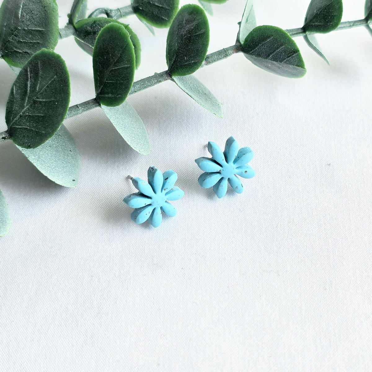 Blue flower-shaped earrings on a white background with green leaves.