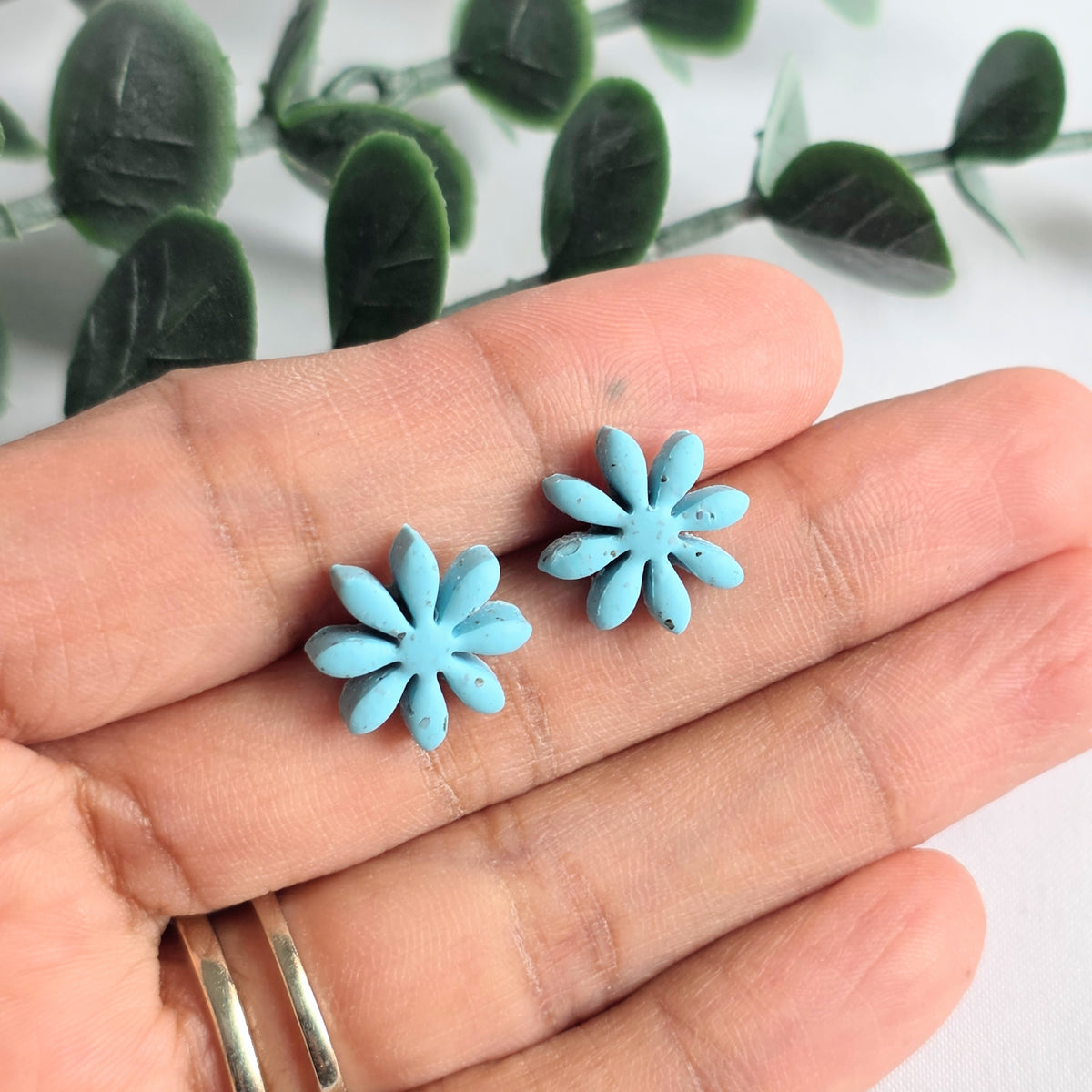 Turquoise flower-shaped earrings held between fingers with a green leafy background