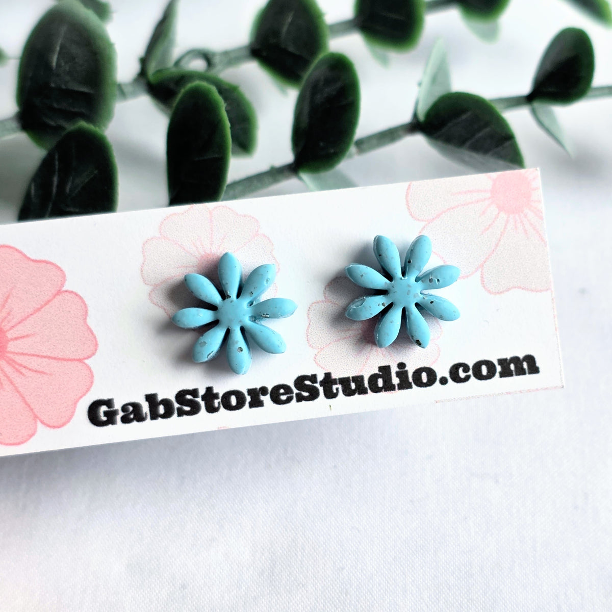 Blue flower-shaped earrings on a card with 'GabStoreStudio.com' branding, against a white background with green leaves.