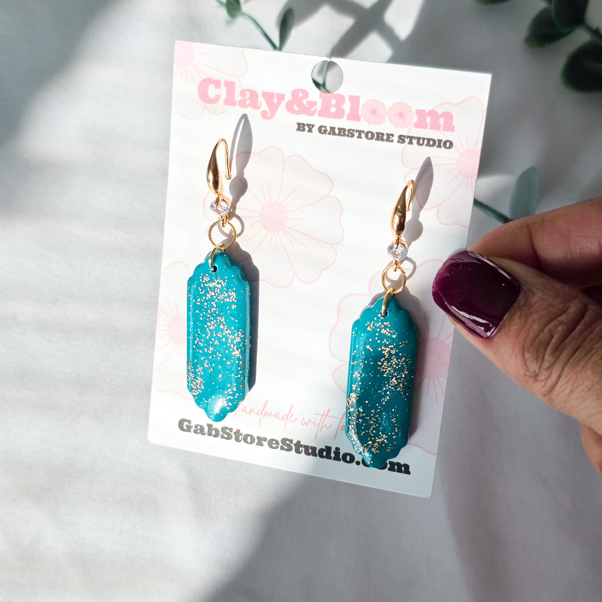 Turquoise earrings with gold accents on a 'Clay & Bloom' branded card held by a hand.