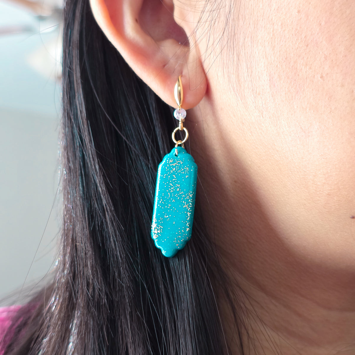 Close-up of a turquoise earring with gold accents worn by a person.