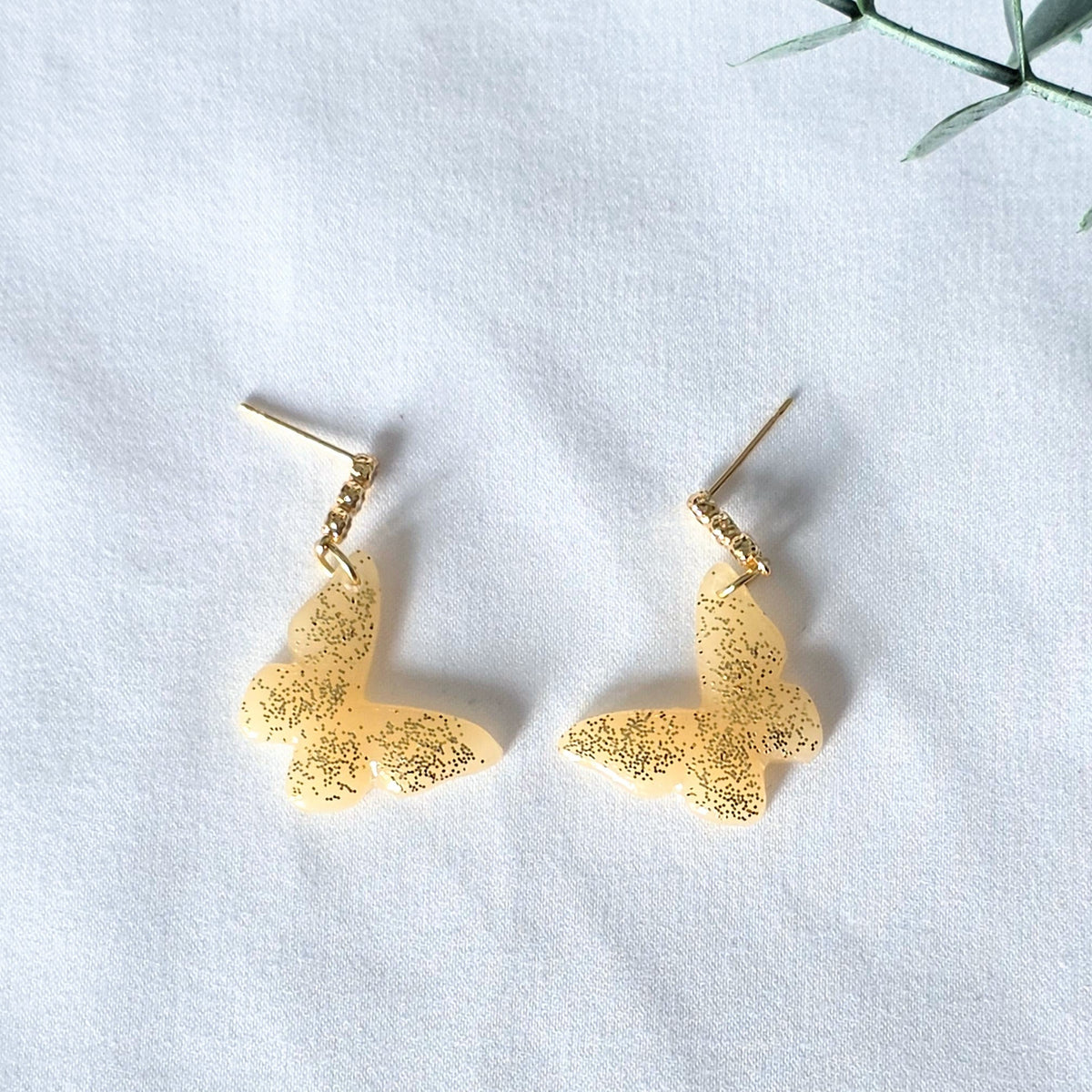 Gold earrings on a white surface with a green leafy branch in the background