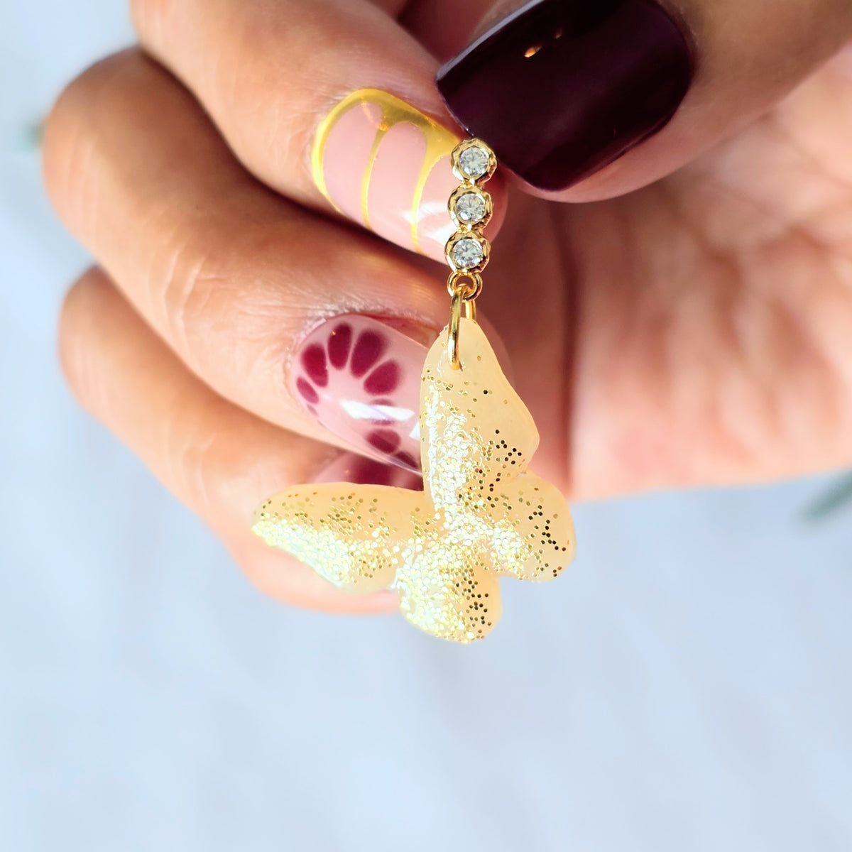 Gold butterfly earring held by a hand with floral nail design on a light background