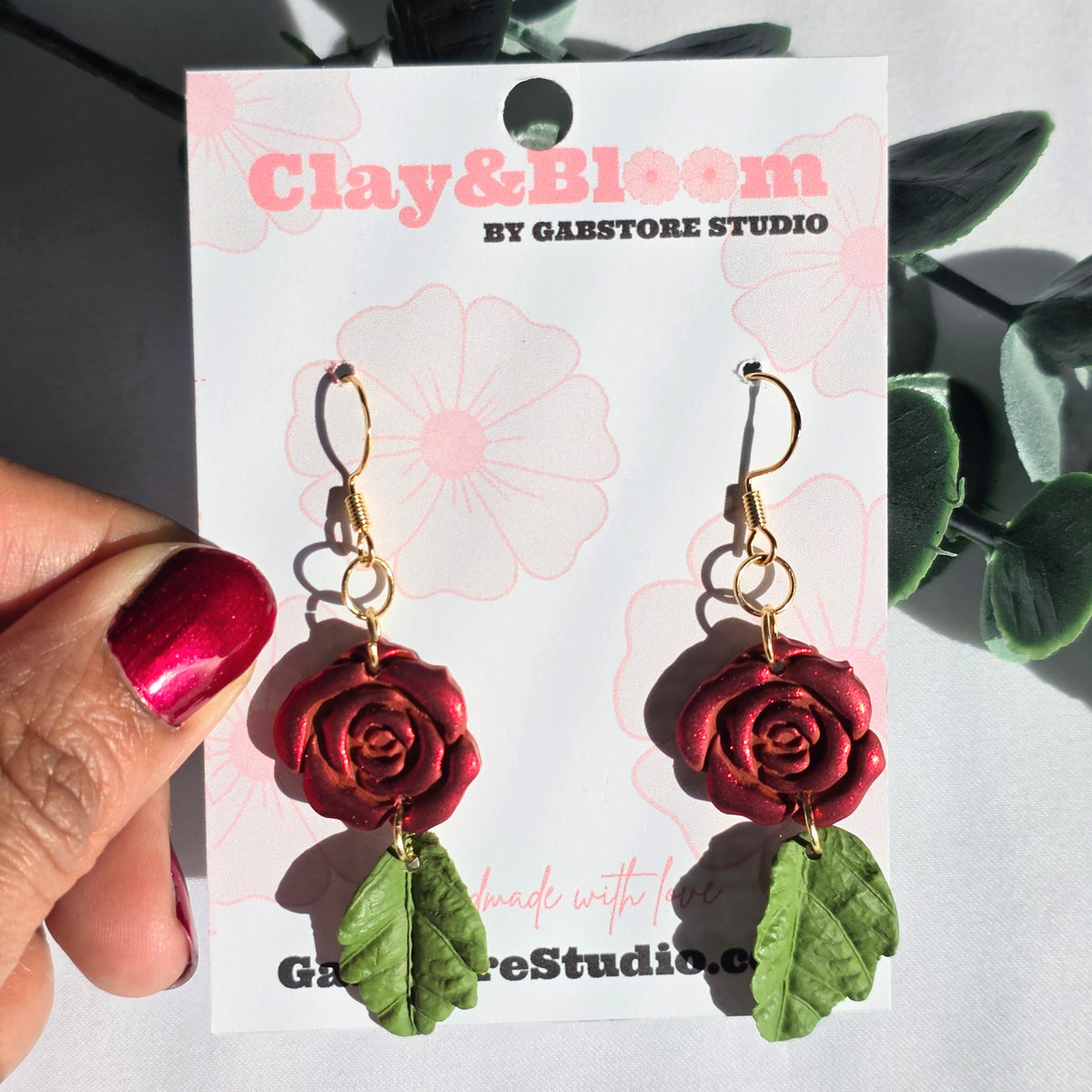 Red rose earrings on a 'Clay & Bloom by Gabstore Studio' card held by a hand.