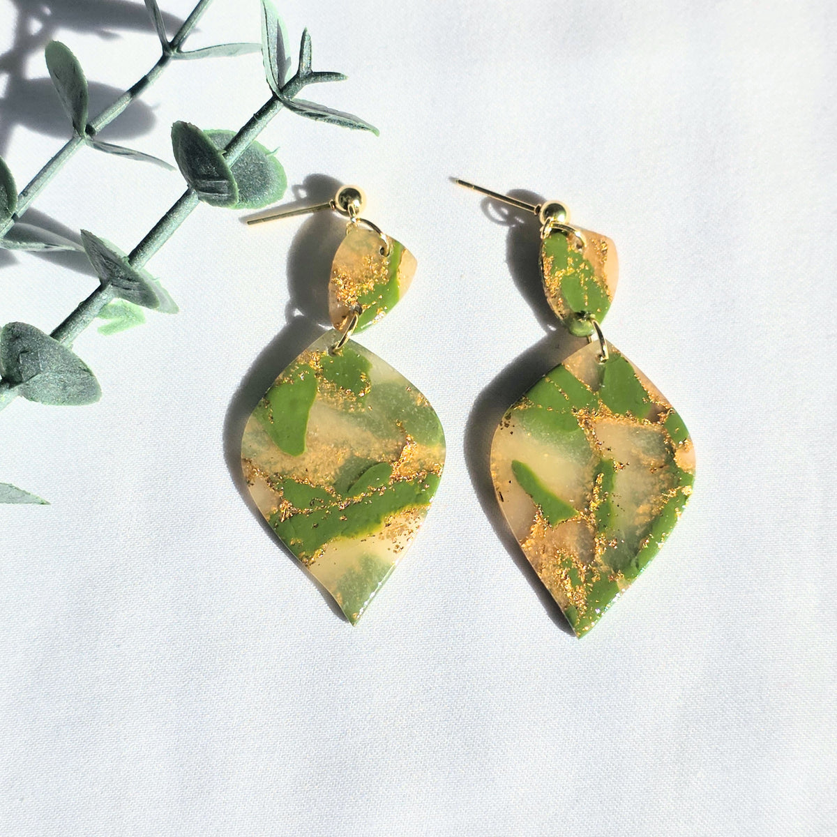 Green and gold leaf-shaped earrings on a white background with green leaves.