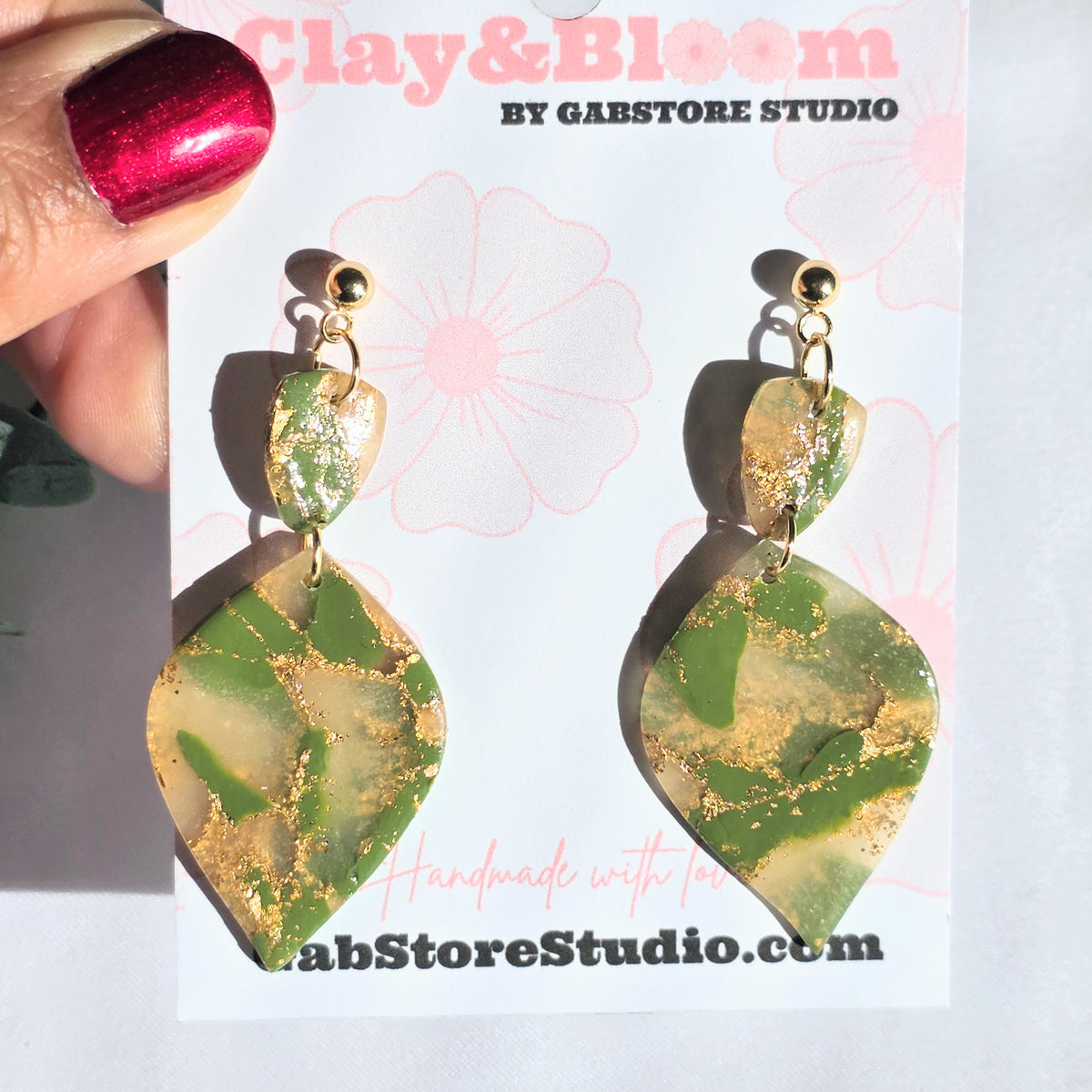 Green and gold earrings held by a hand with red nail polish, displayed on a white card with 'Clay & Bloom' branding.