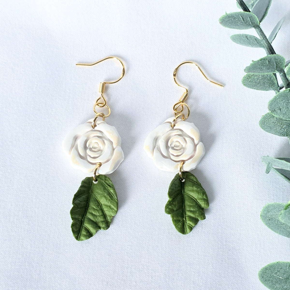 Pair of earrings with white rose and green leaf design on a light background