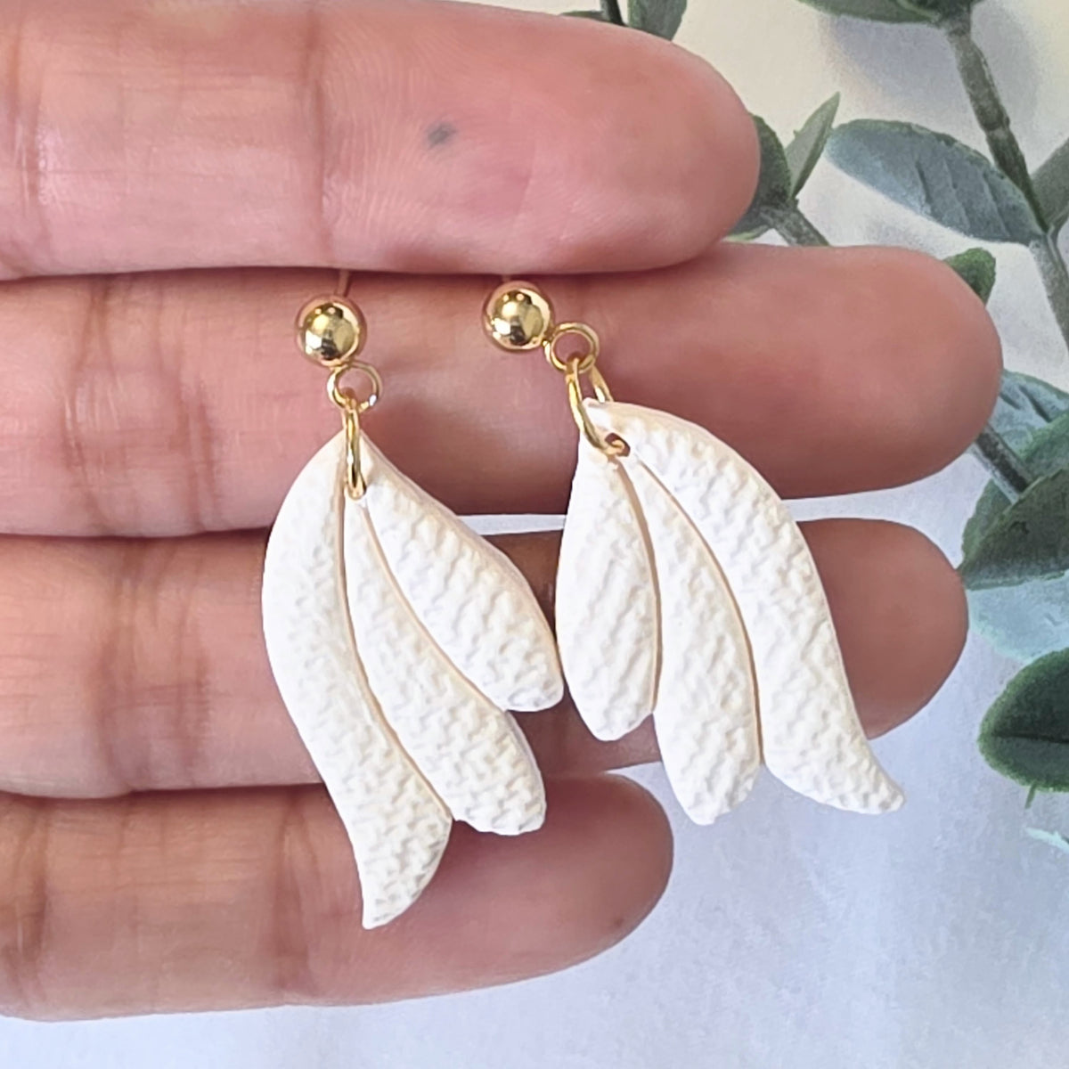 White leaf-shaped earrings with gold hooks held between fingers against a light background