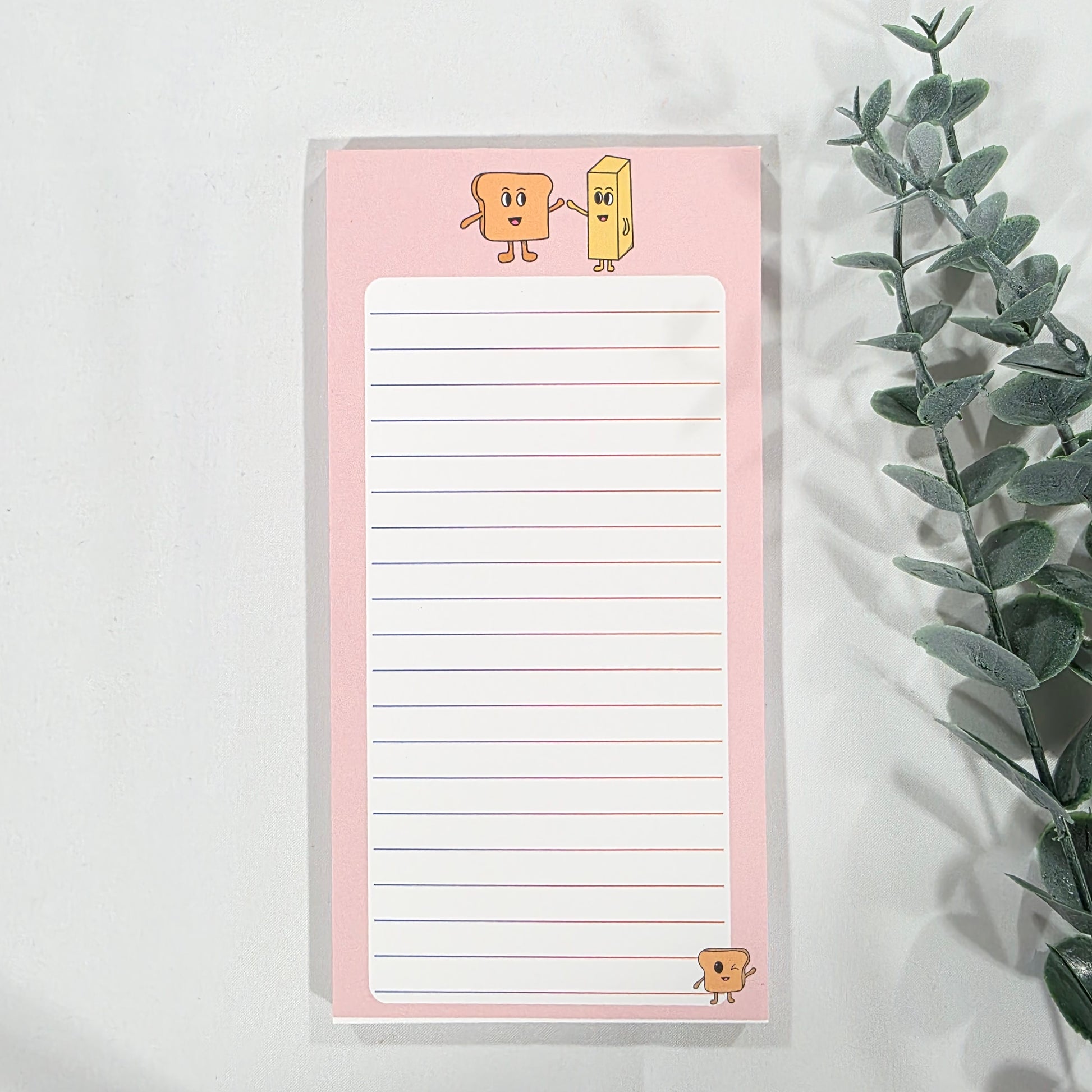 Notepad with lined paper on a pink border, featuring cartoon toast and butter illustrations, on a white background with eucalyptus leaves.