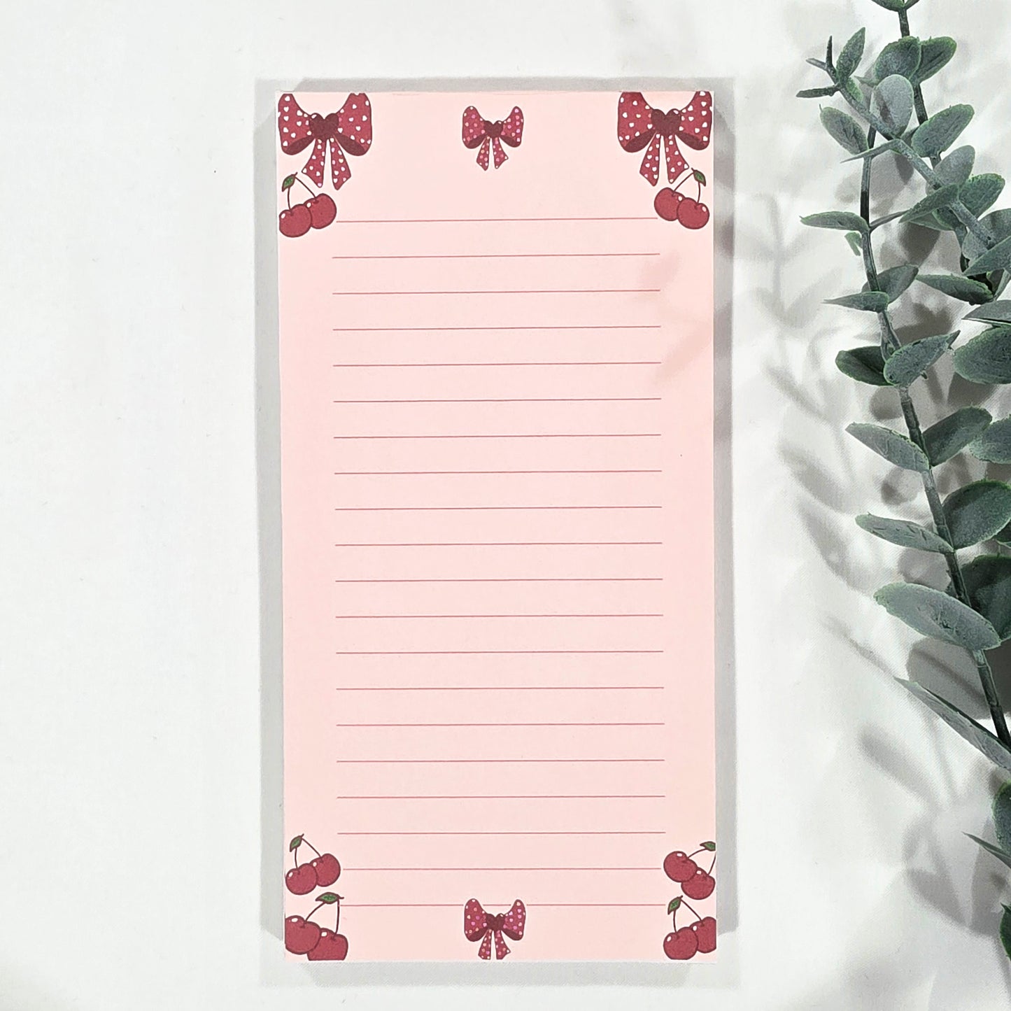 Pink notepad with cherry and bow designs on a white surface with a plant.
