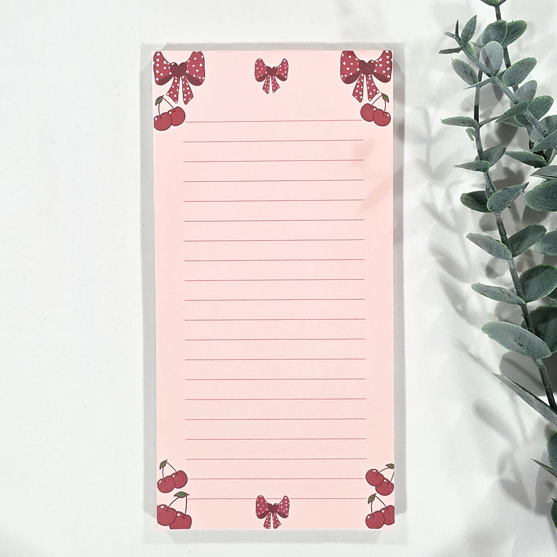 Pink notepad with cherry and bow designs on a white surface with a plant.