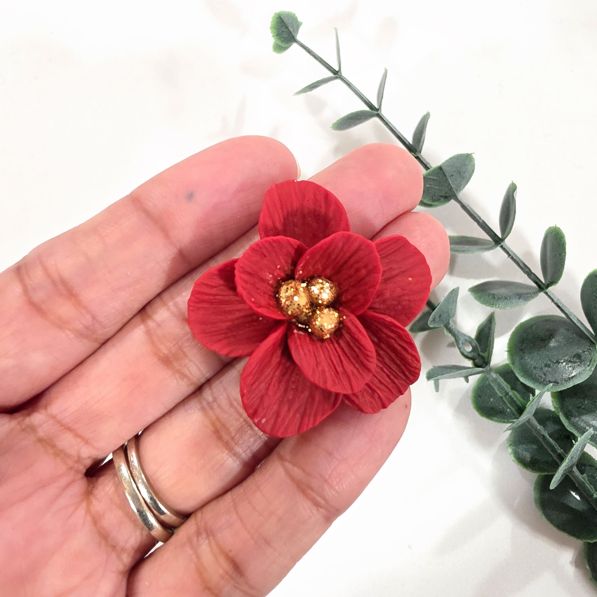 Red flower-shaped magnet made from polymer clay held in a hand with green leaves in the background.