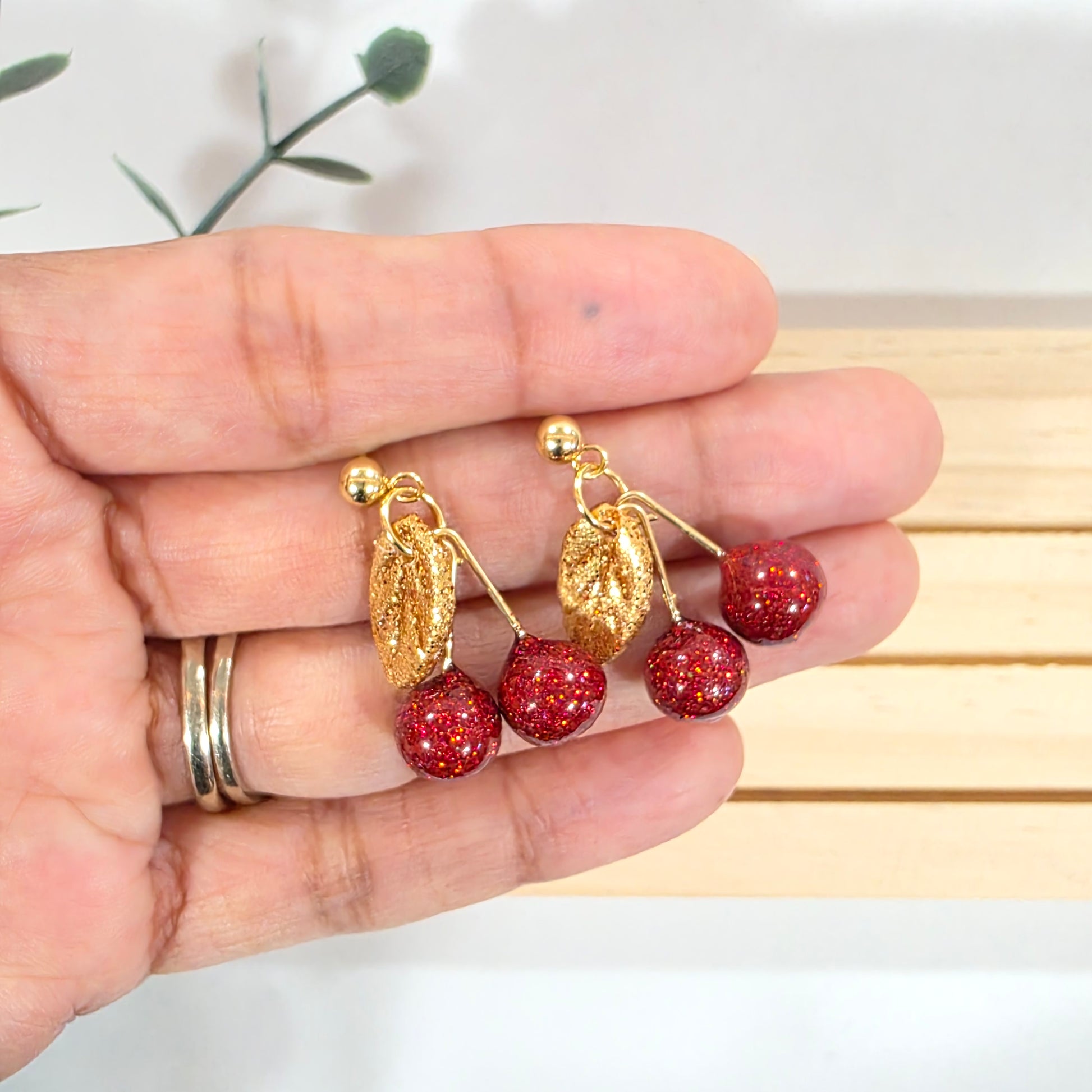 Hand holding a pair of earrings with red polymer clay cherries and gold accents against a light background.