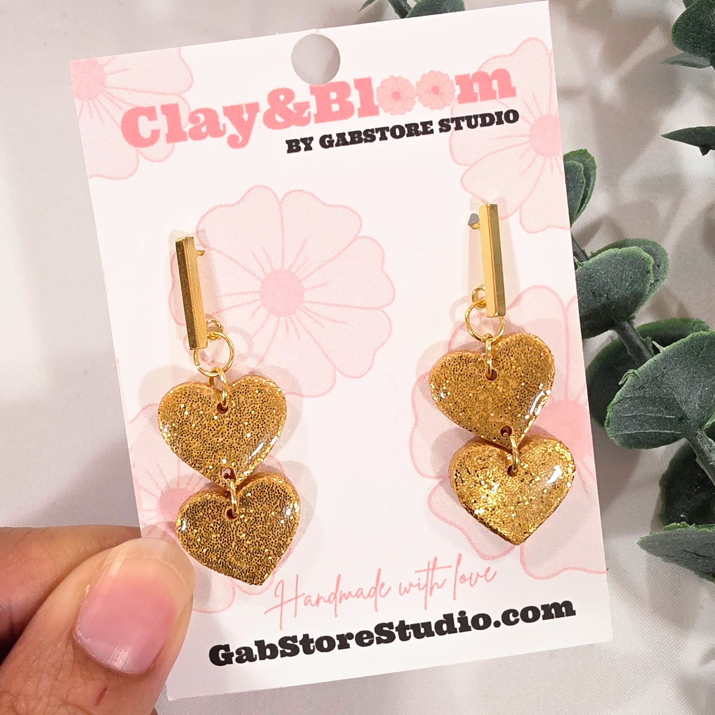 Gold heart-shaped earrings on a Clay&Bloom branded card with a hand holding it.