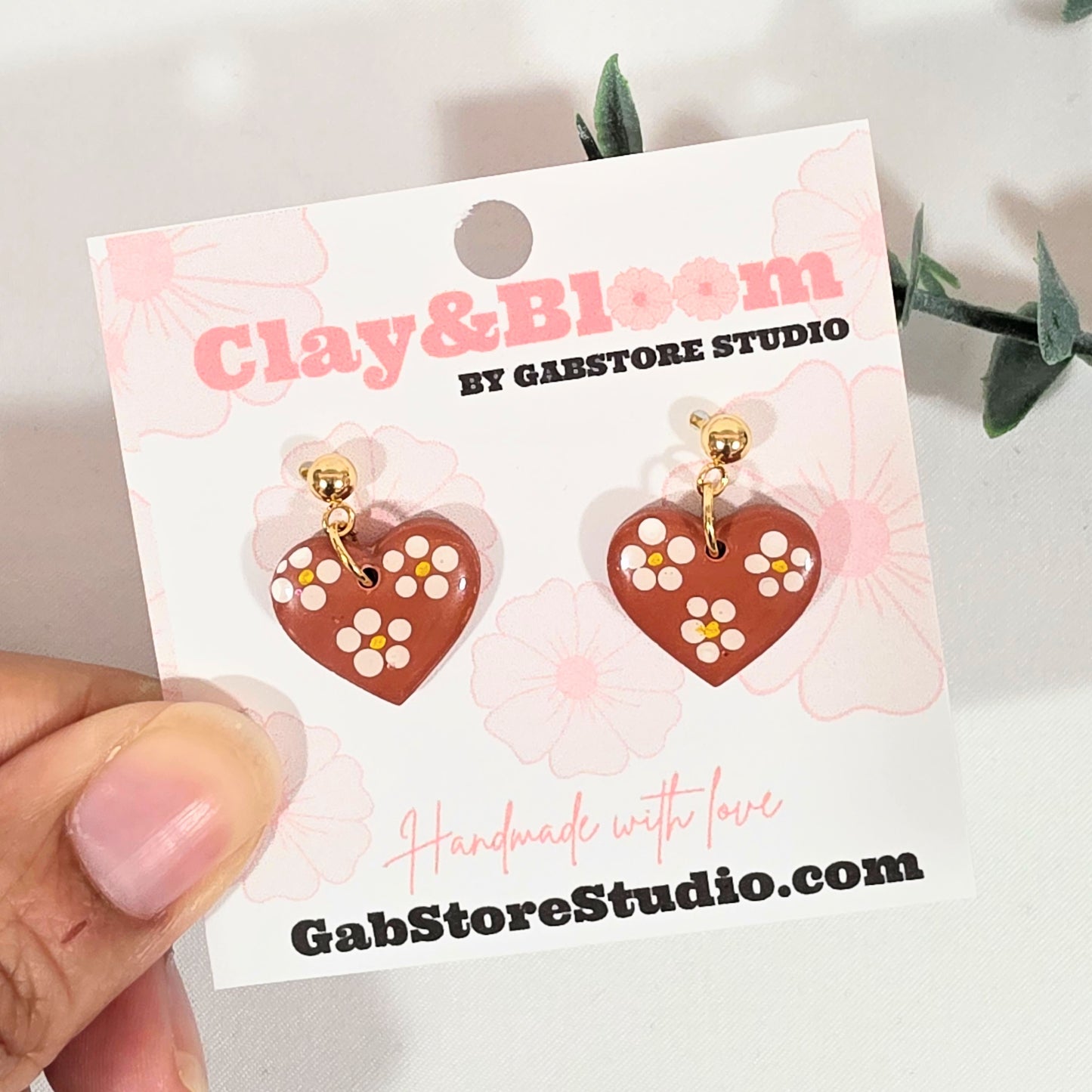 Heart-shaped earrings on a 'Clay & Bloom' branded card held by a hand.