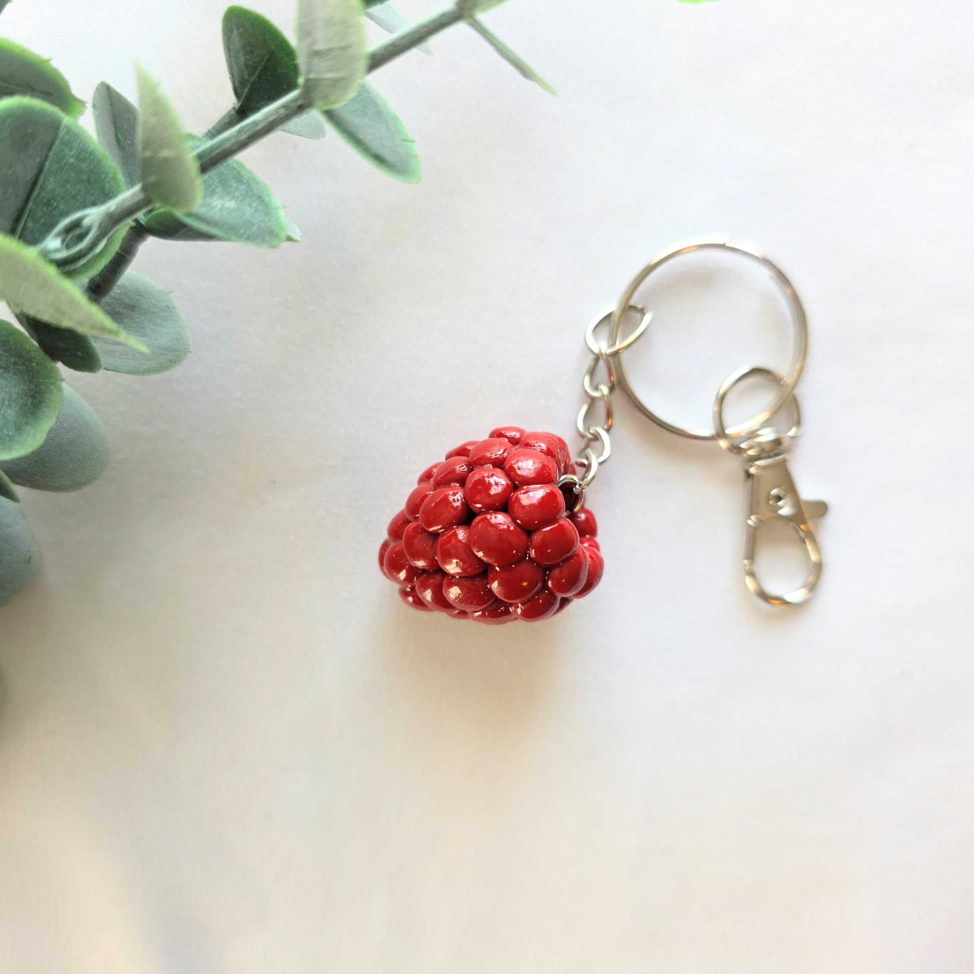 Red raspberry made from polymer clay, keychain. Sitting on a white background and greenery.
