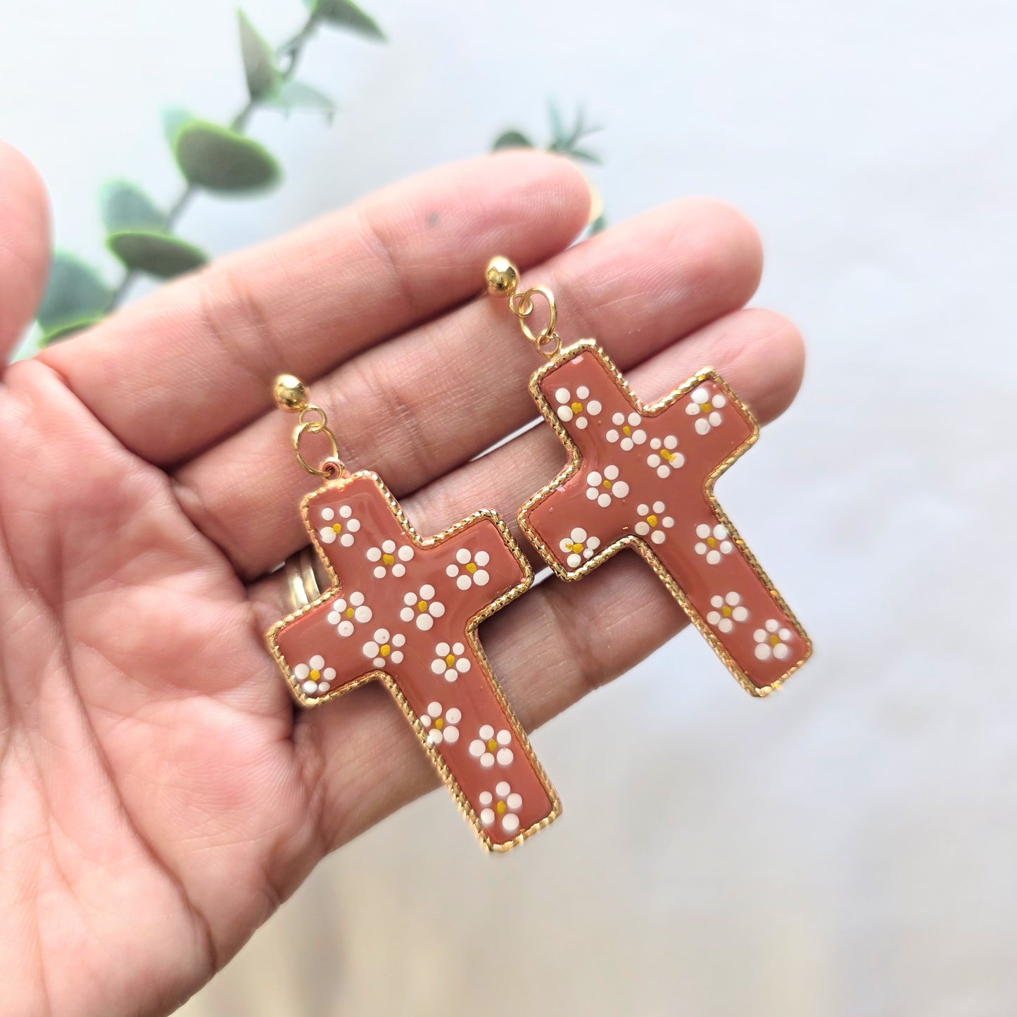 Handmade Mexican pottery–inspired cross earrings with gold plated brass, terracotta-colored polymer clay center, hand-painted white floral details, and 24k gold plated ball stud posts, on a hand on a neutral background.