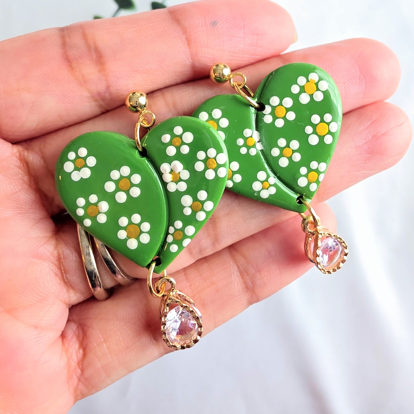 Green heart-shaped earrings with floral patterns held in a hand against a light background