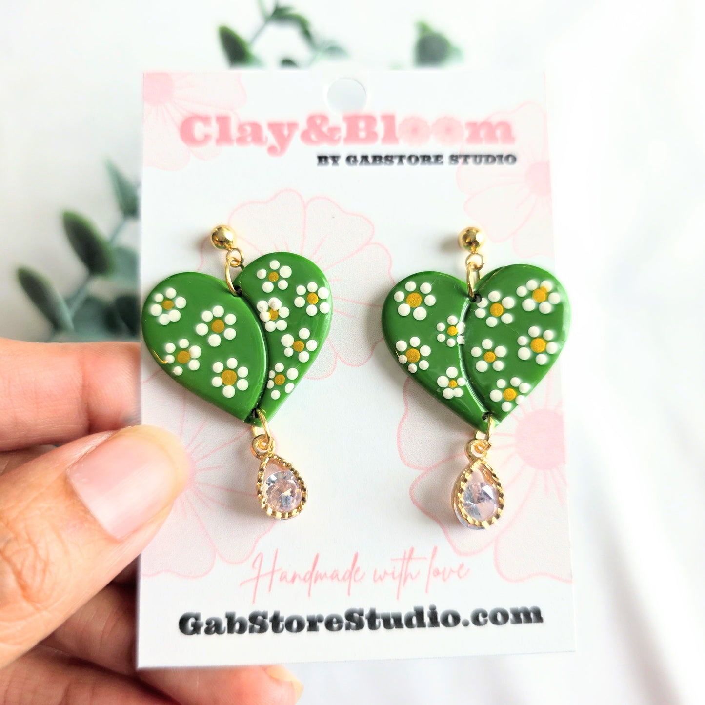 Green heart-shaped earrings with floral designs on a 'Clay & Bloom' branded card.