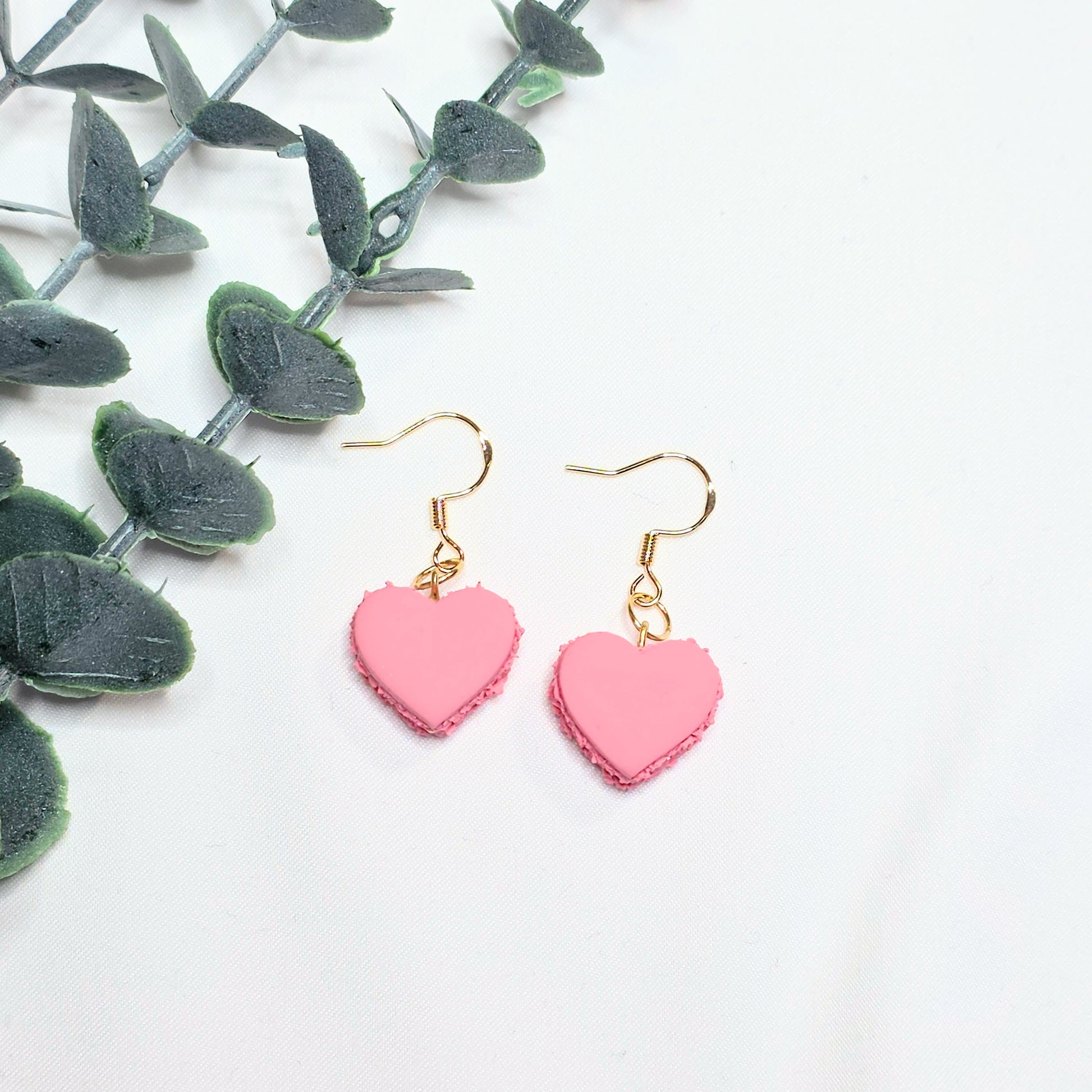Pink heart-shaped earrings on a white background with green leaves.