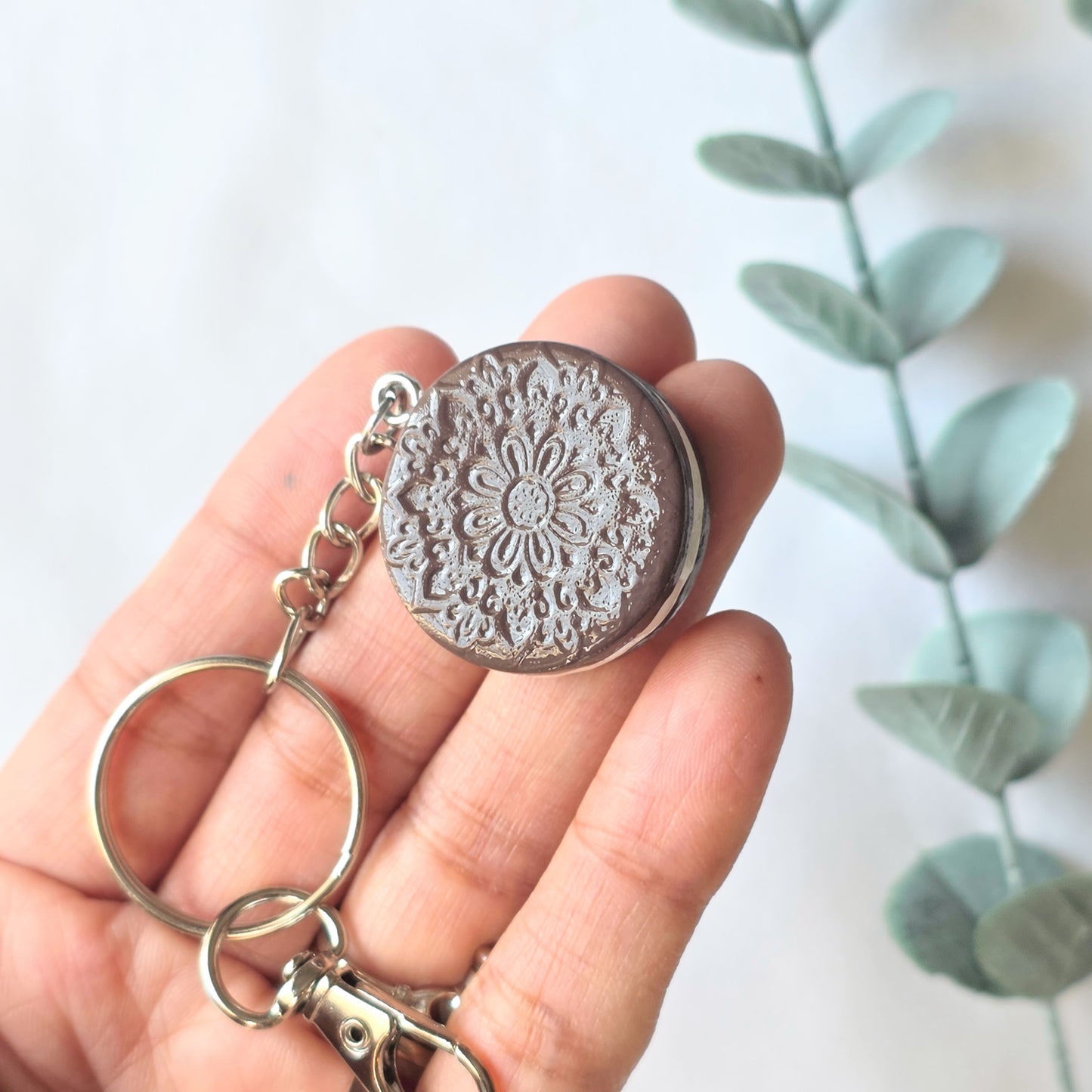 Hand holding a silver keychain of cookie with intricate design against a light background