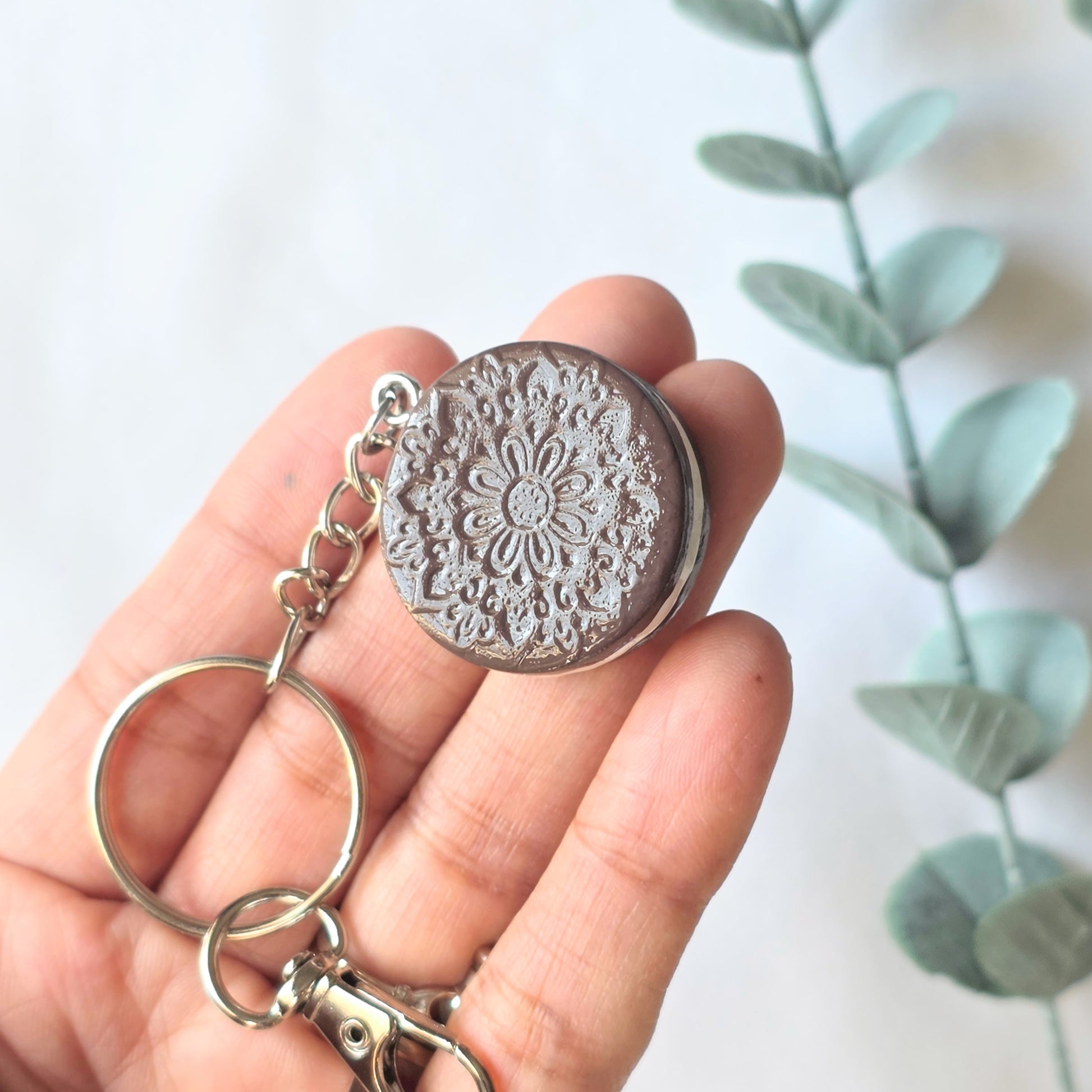 Hand holding a silver keychain of cookie with intricate design against a light background