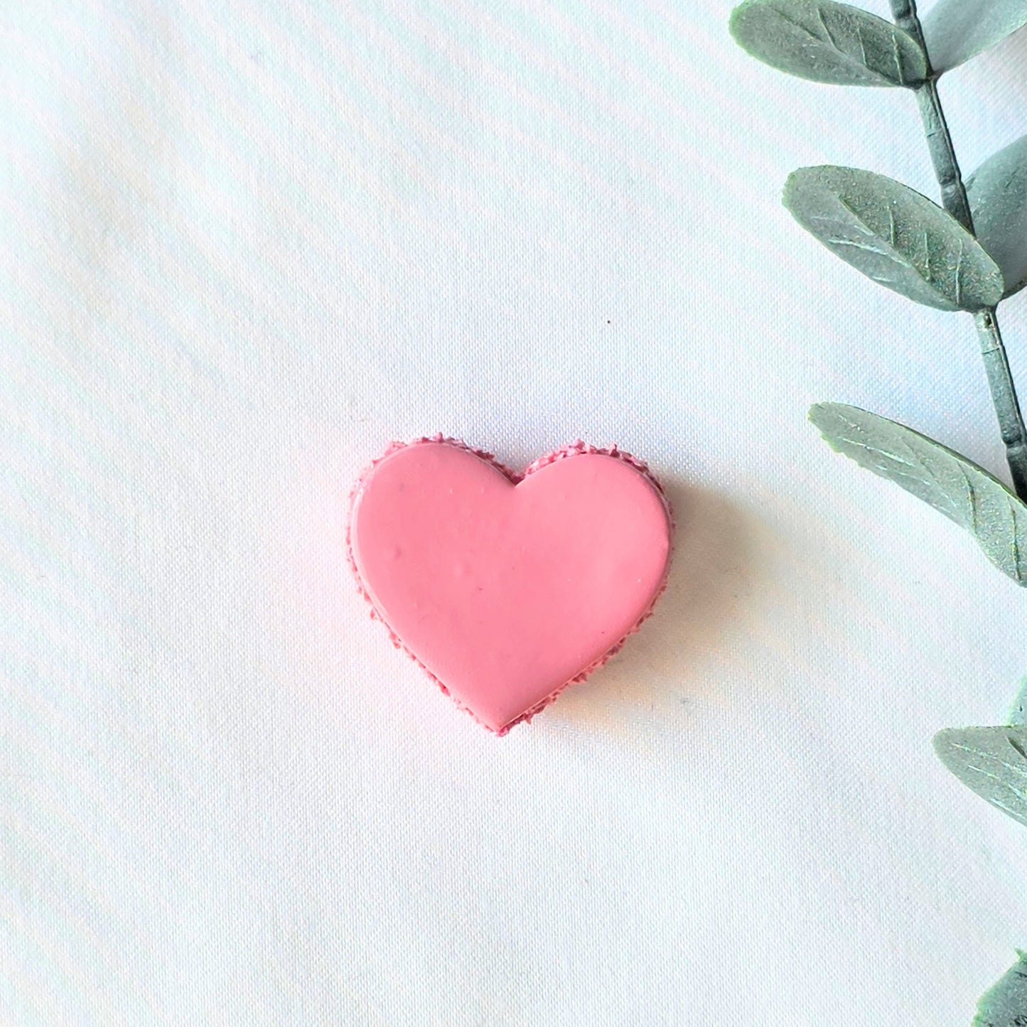 Pink heart-shaped macaron on a white background with a green leafy branch.