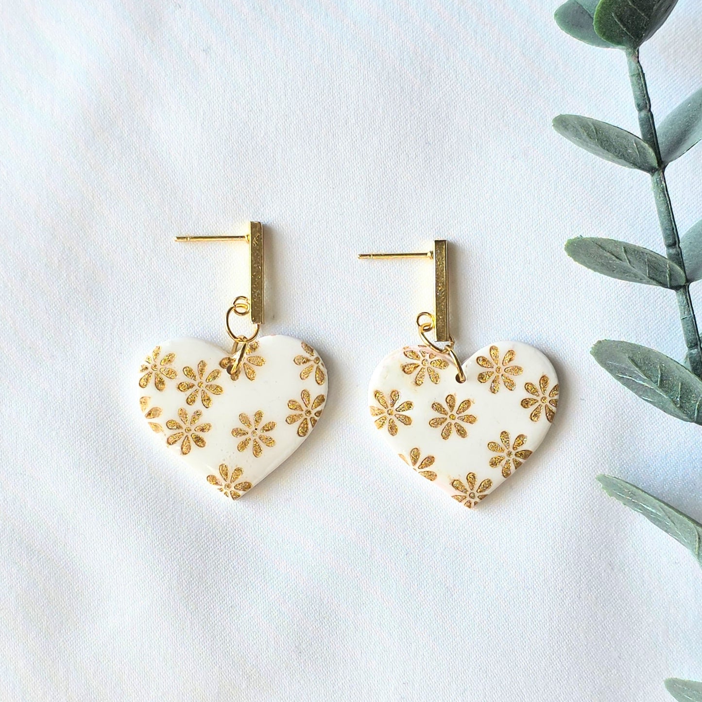 Heart-shaped earrings with gold floral patterns on a white background with green leaves.