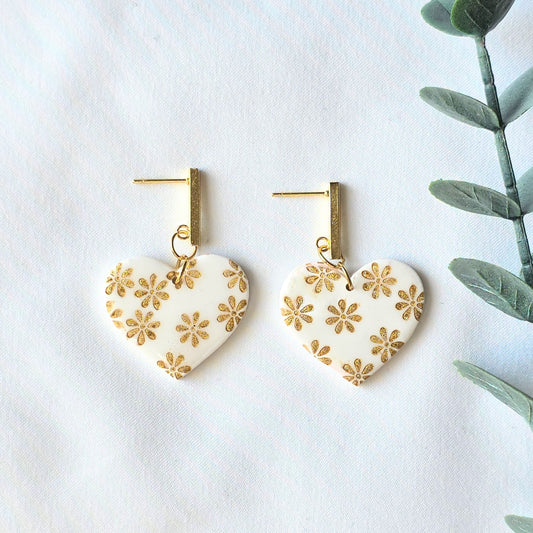 Heart-shaped earrings with gold floral patterns on a white background with green leaves.