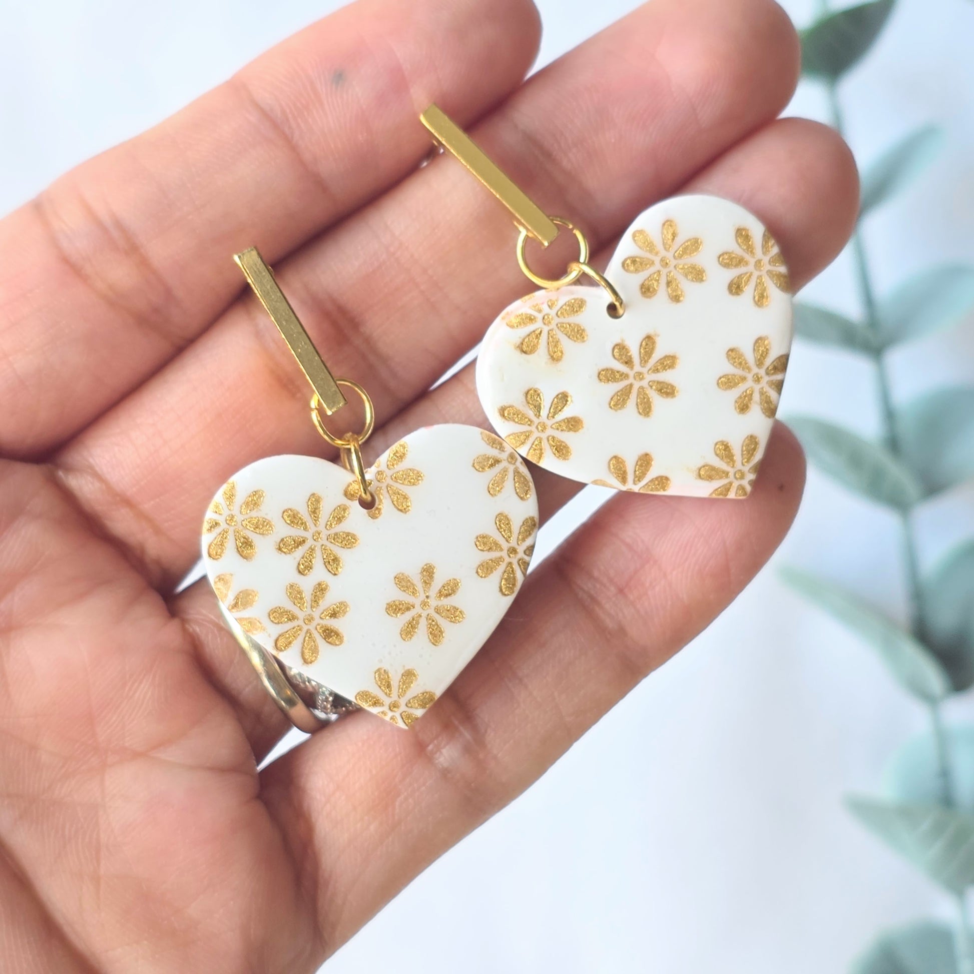 Heart-shaped earrings with gold floral patterns held in a hand against a blurred natural background