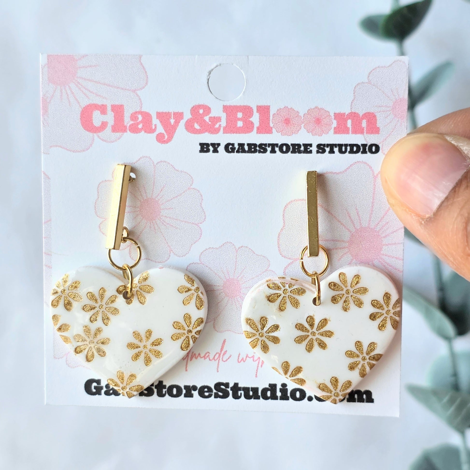 Heart-shaped earrings with gold floral patterns on a 'Clay & Bloom' branded card.