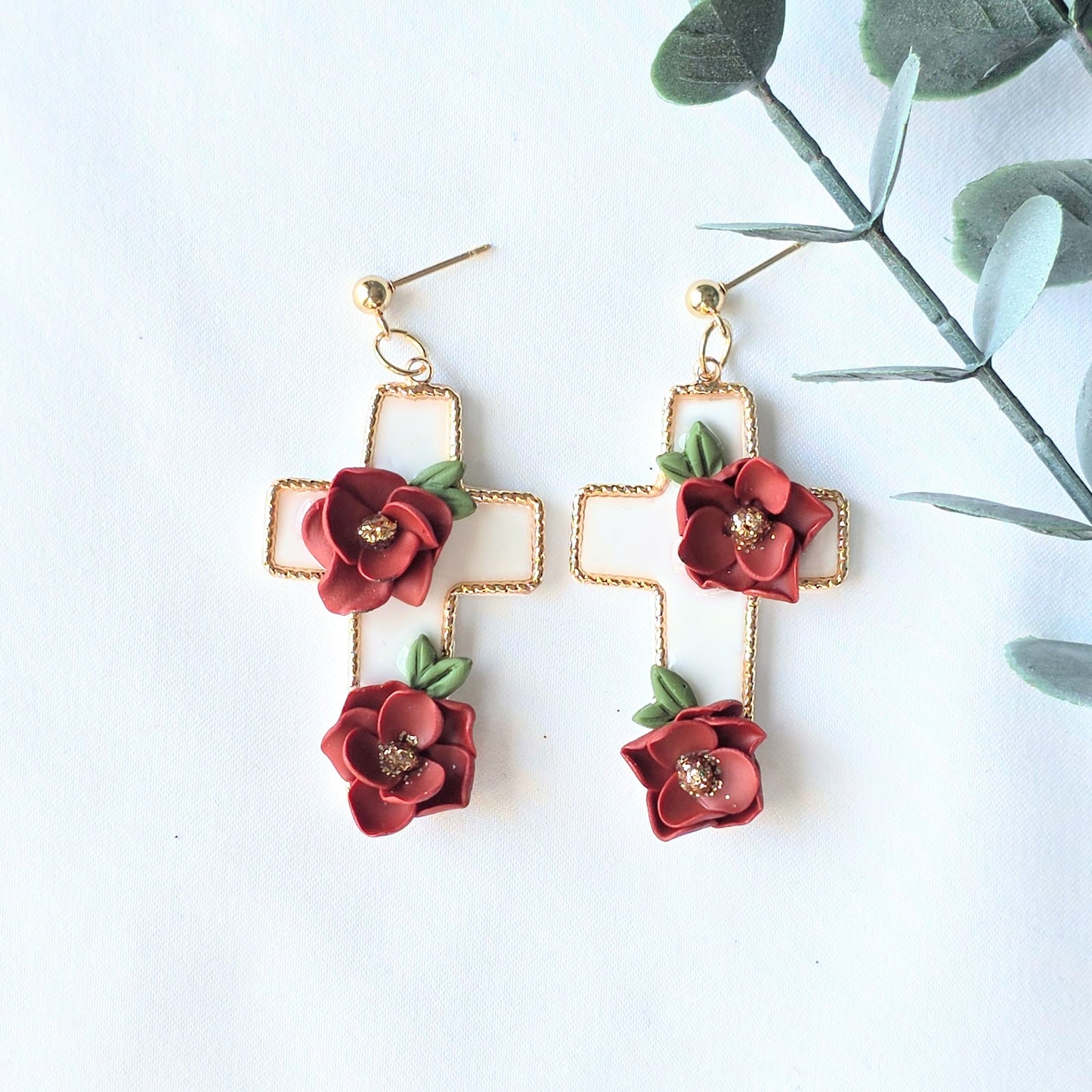 Gold cross-shaped earrings with red floral elements on a white background