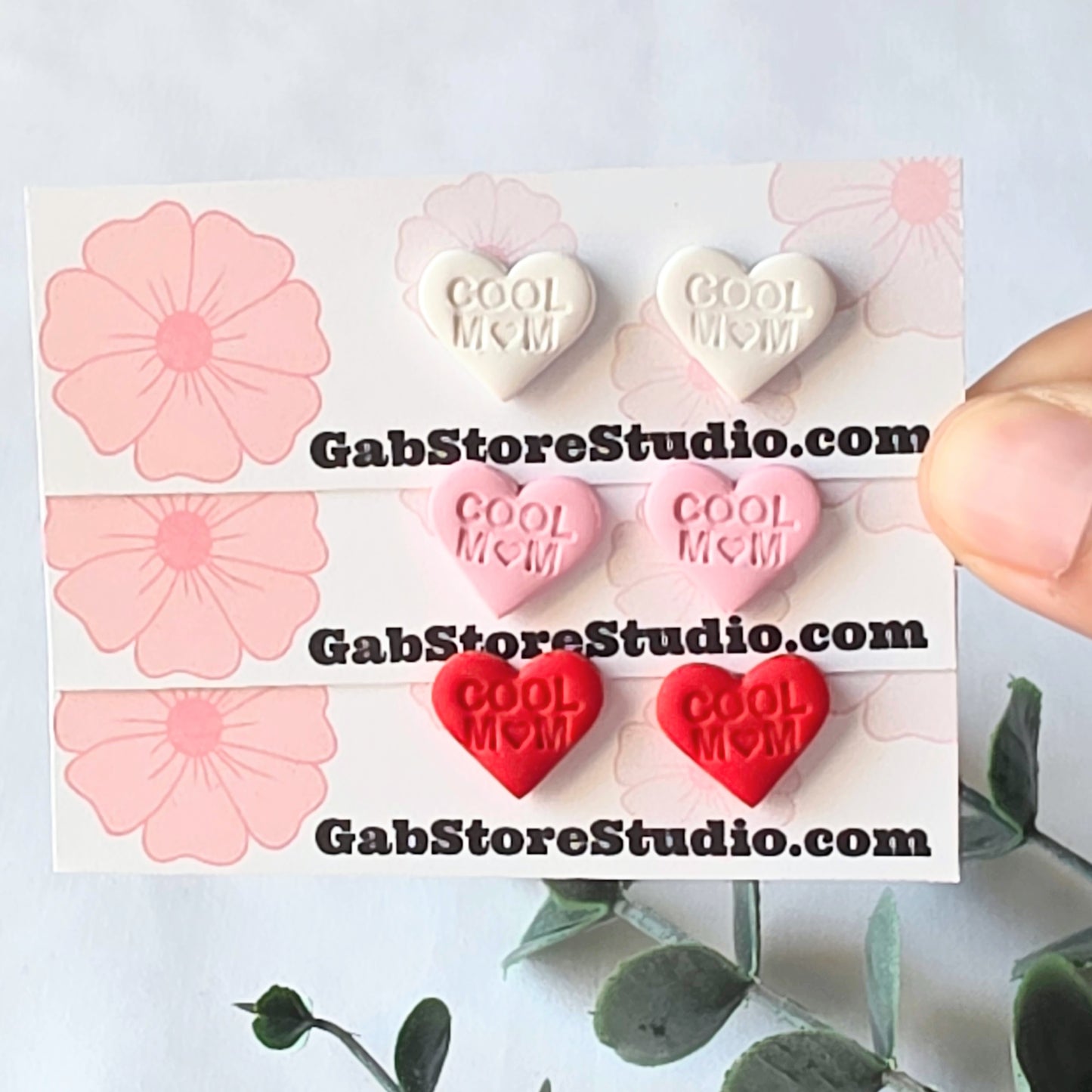 Heart-shaped earrings with 'Cool Mom' text on a card with floral designs, held by a hand.