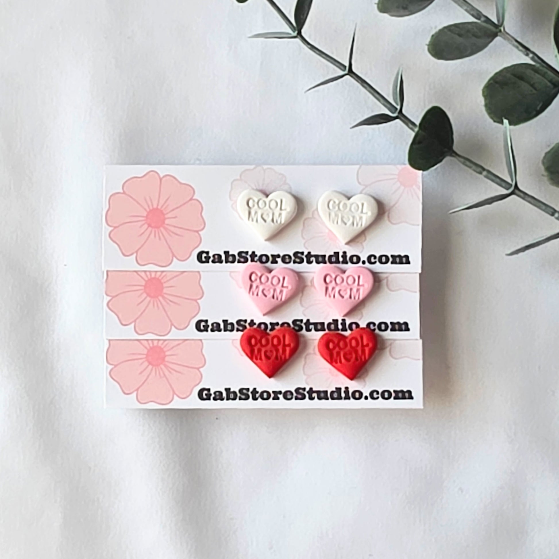 Heart-shaped earrings with 'Cool Mom' text on a branded card, set against a white background with floral elements.