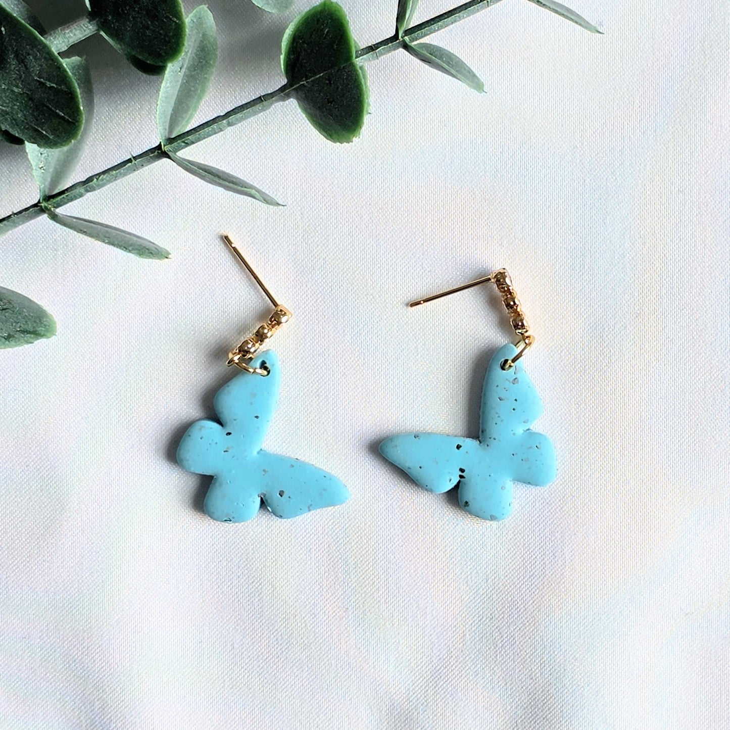 Turquoise butterfly earrings on a white background with green leaves.