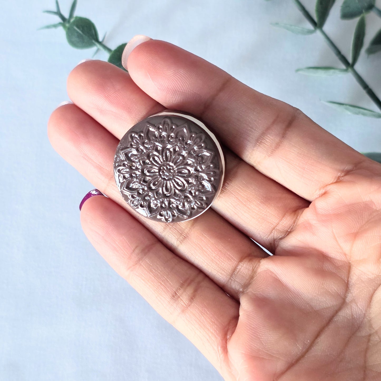 Hand holding a polymer clay floral-patterned cookie against a white background with green leaves.