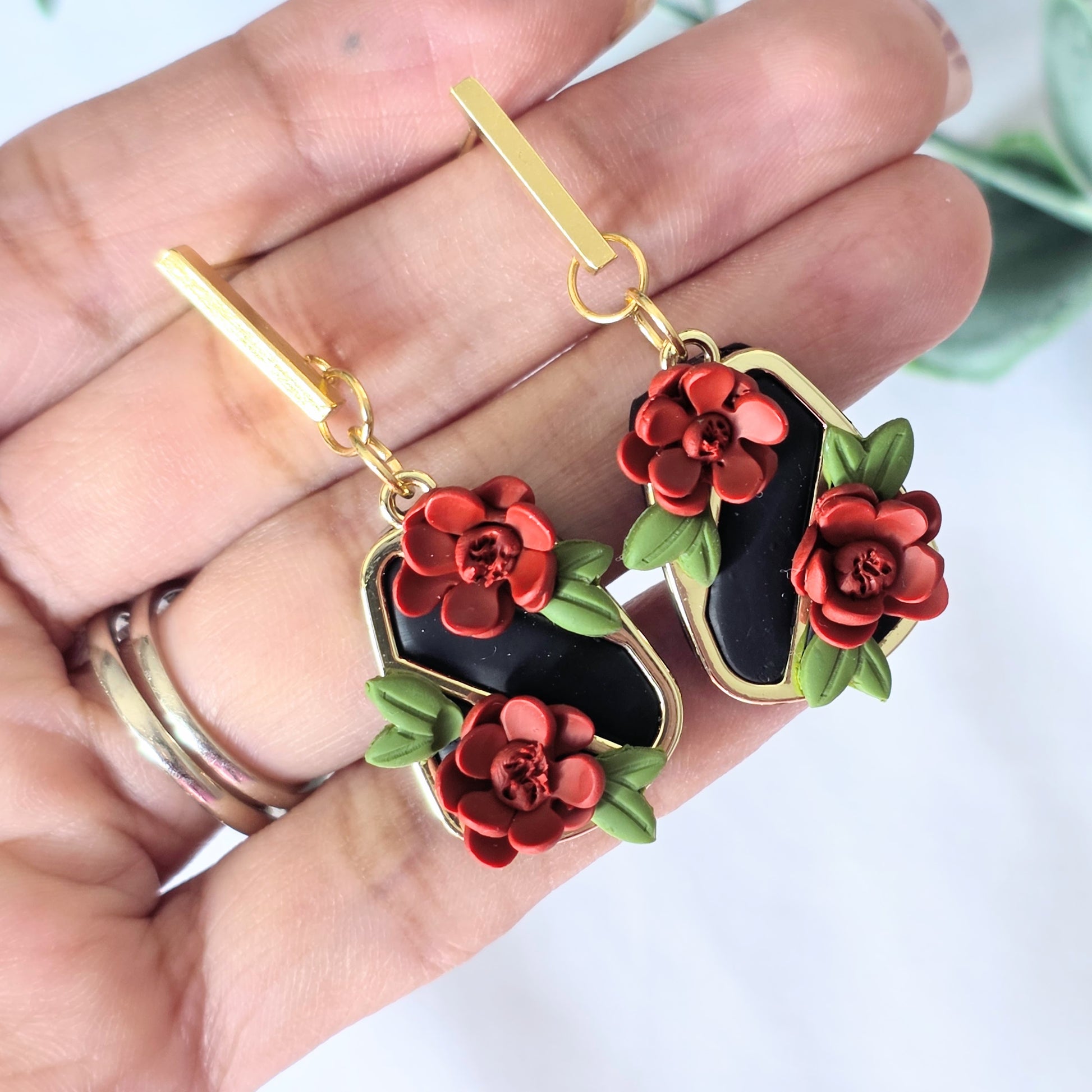 Gold earrings with red floral designs held in a hand against a white background