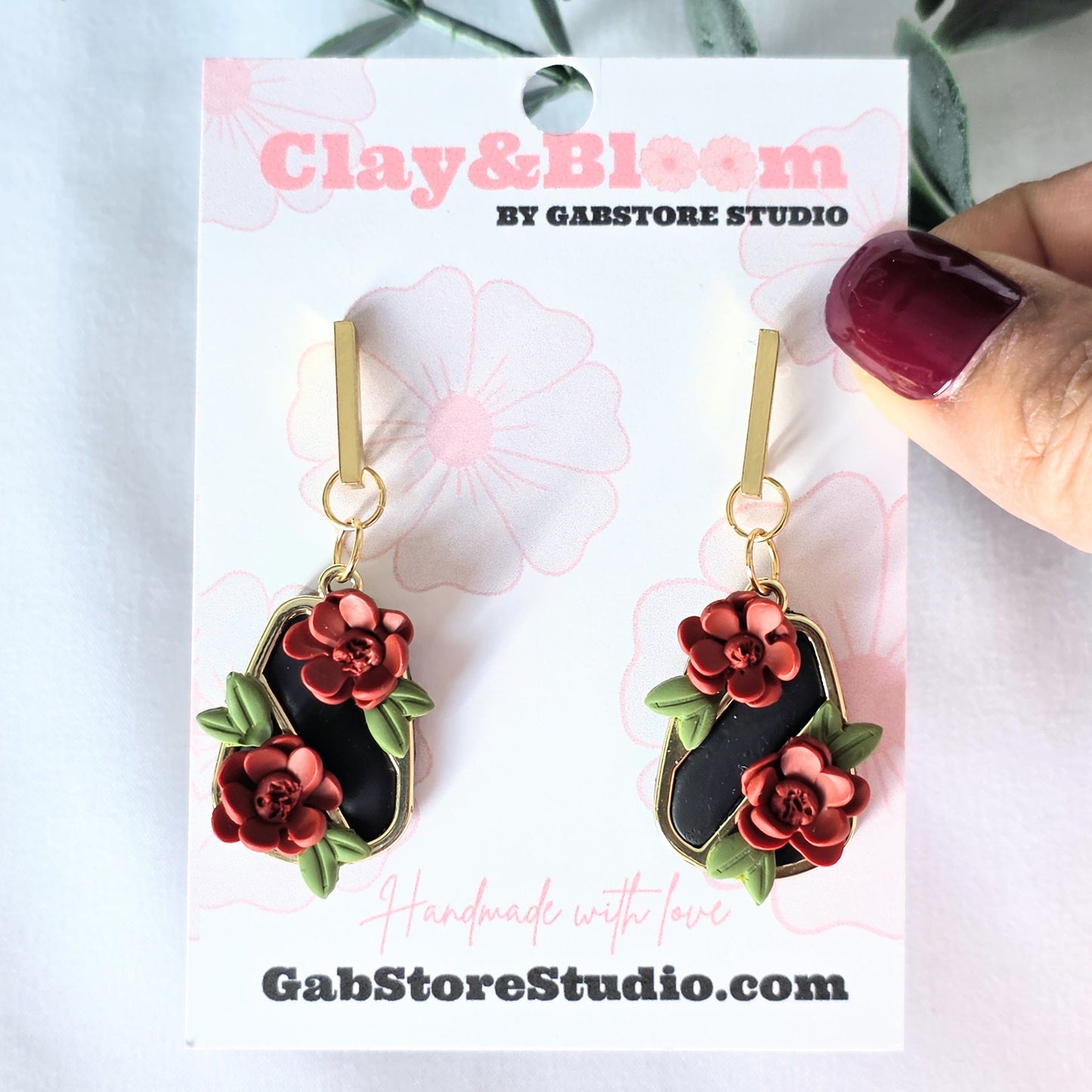 Floral earrings on a Clay&Bloom branded card with a hand holding one of the earrings.