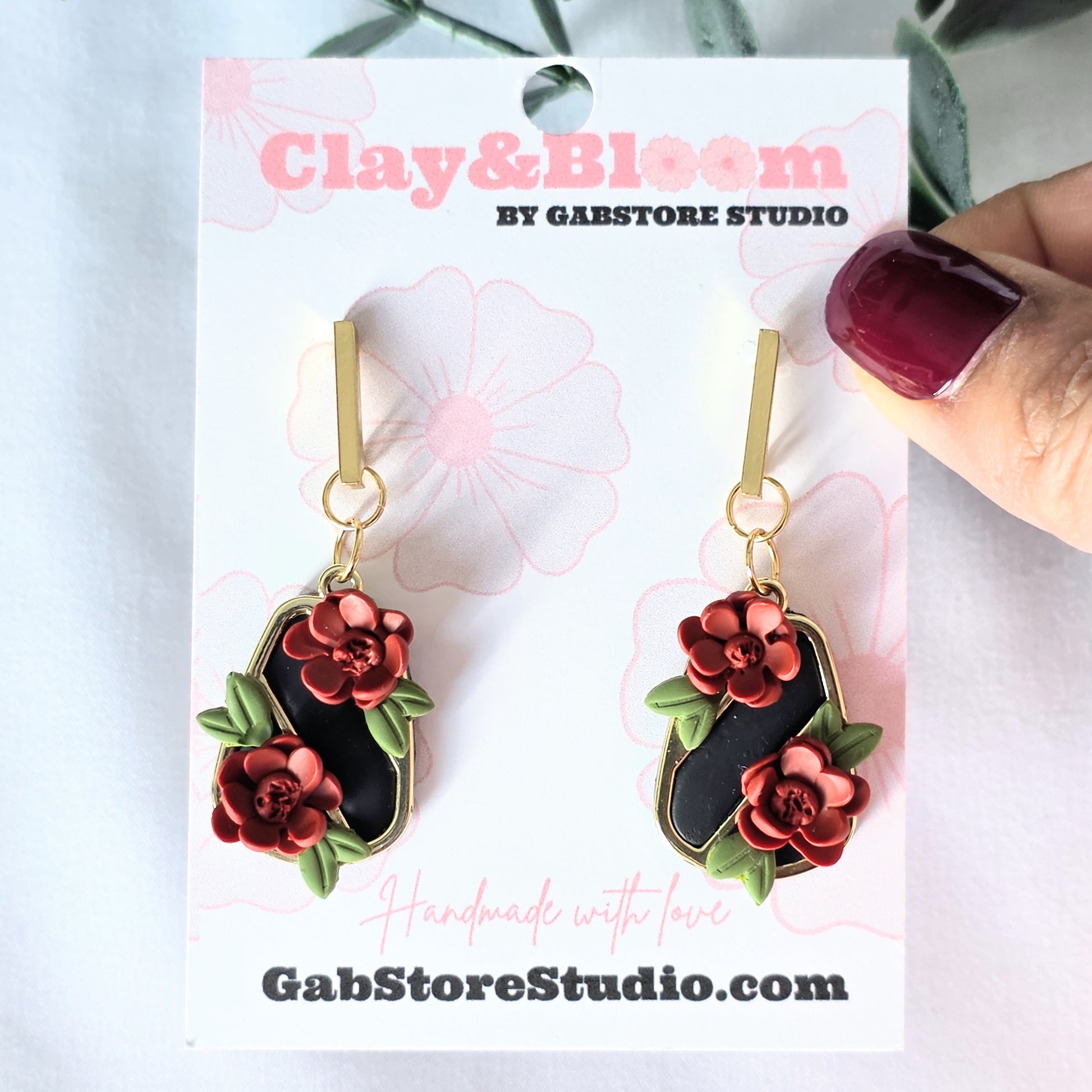 Floral earrings on a Clay&Bloom branded card with a hand holding one of the earrings.