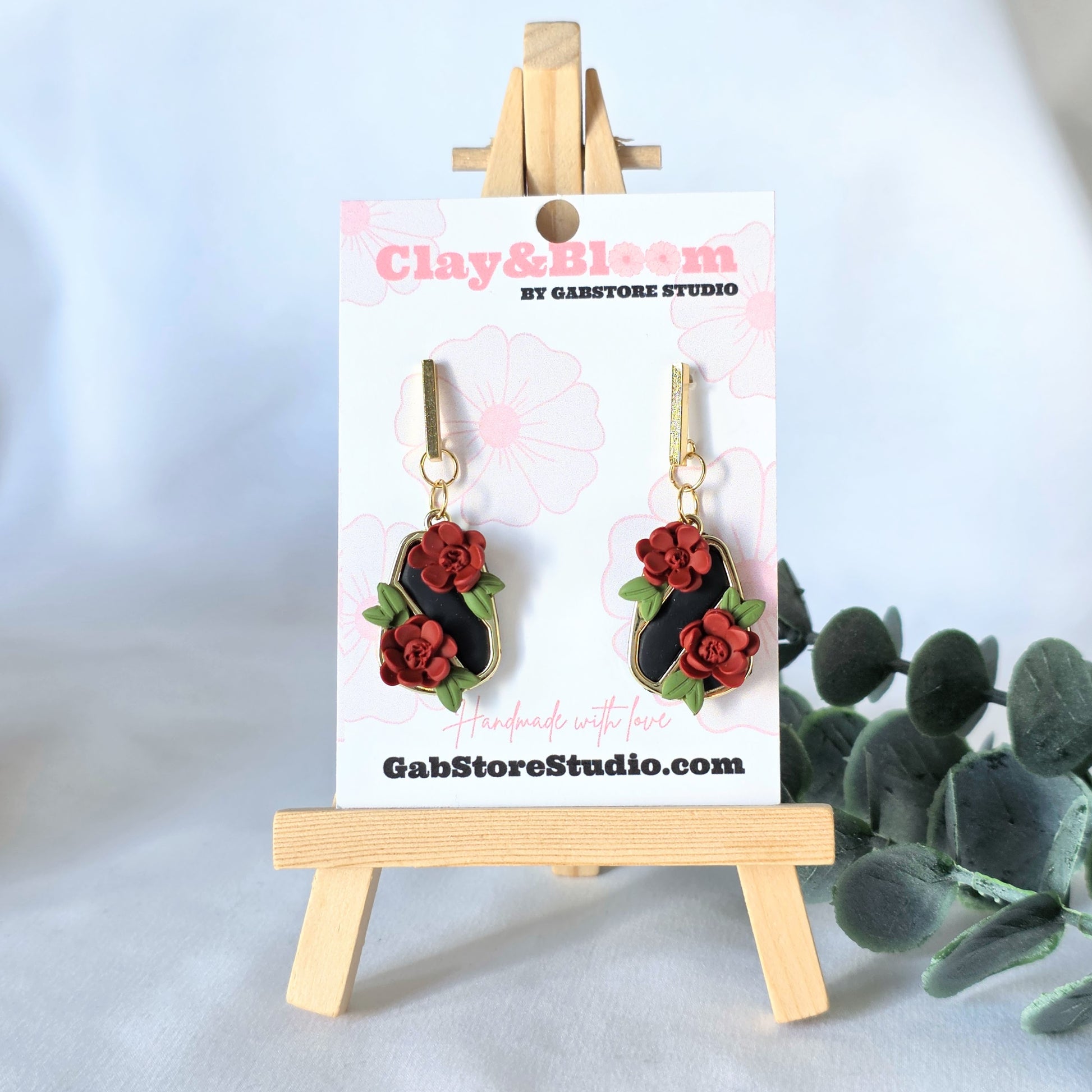 Floral earrings on a display card with 'Clay & Bloom' branding, placed on a light blue background.