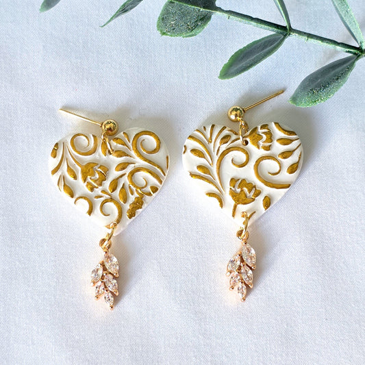 Heart-shaped earrings with gold filigree design on a white background with green leaves.